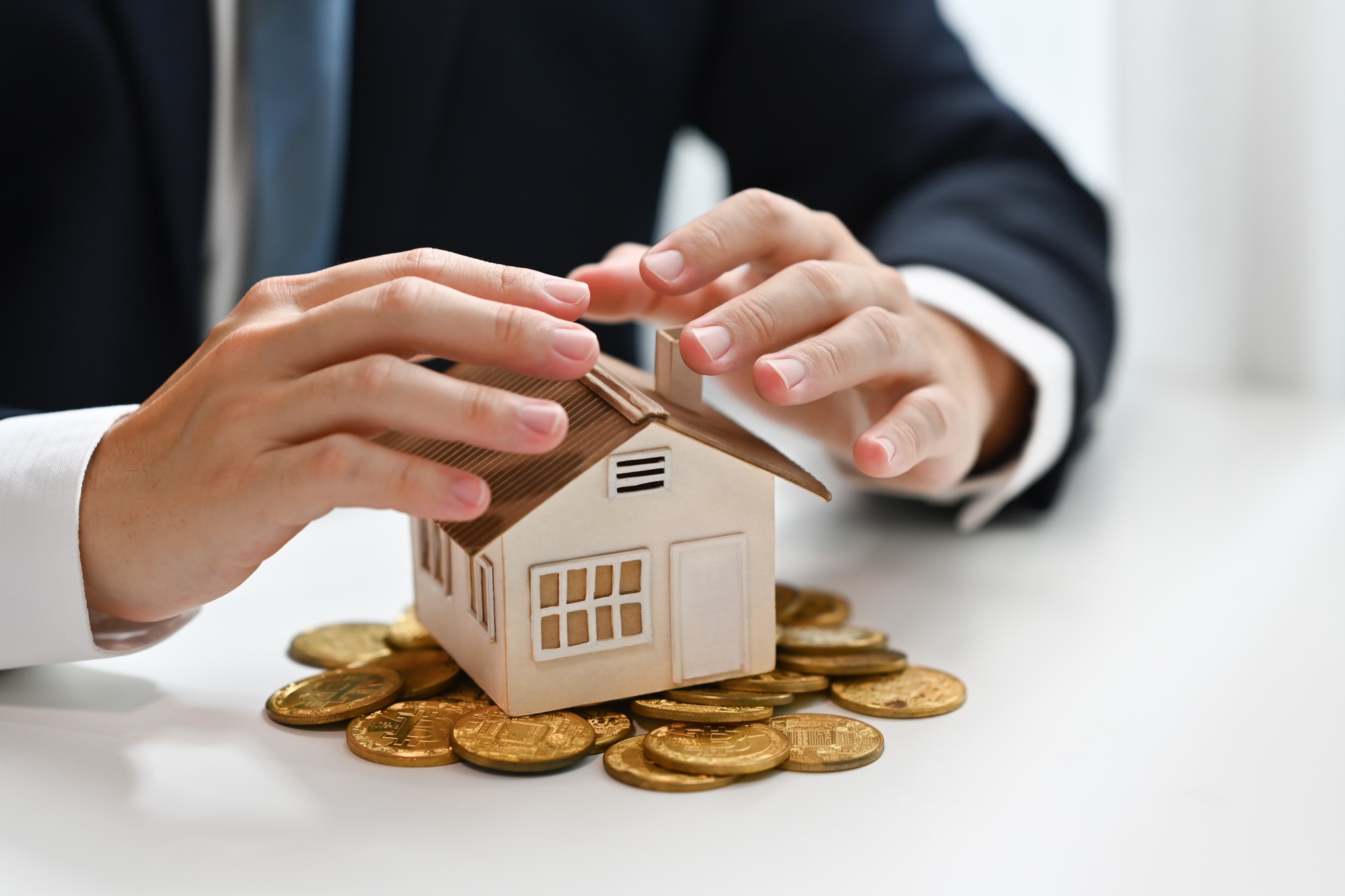 Businessman hands protecting house model and pile of gold coins.