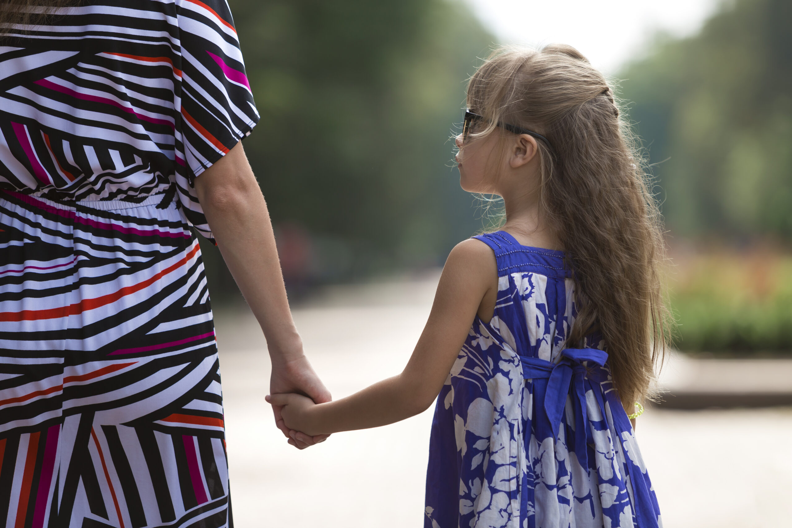 Back view of young blond long-haired attractive woman and small child girl holding hands outdoors.